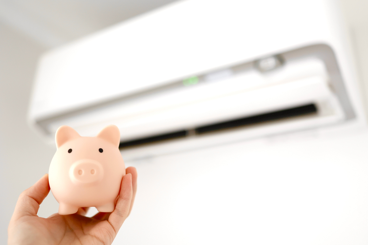 A person holds a piggy bank beneath an air conditioner, representing ways to reduce cooling costs in Virginia.